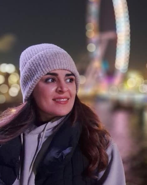 Parisa Hosseini smiling and looking to the side, in a light grey knitted hat, by the water at night with a Ferris wheel and city lights blurred behind her.